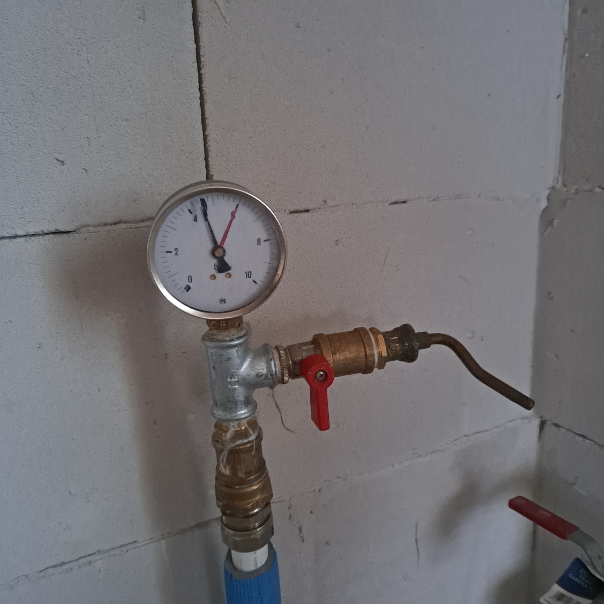 Zamknięty zawór z manometrem na tle ściany z betonu komórkowego. Widoczne elementy instalacji hydraulicznej. Czerwony uchwyt zaworu.