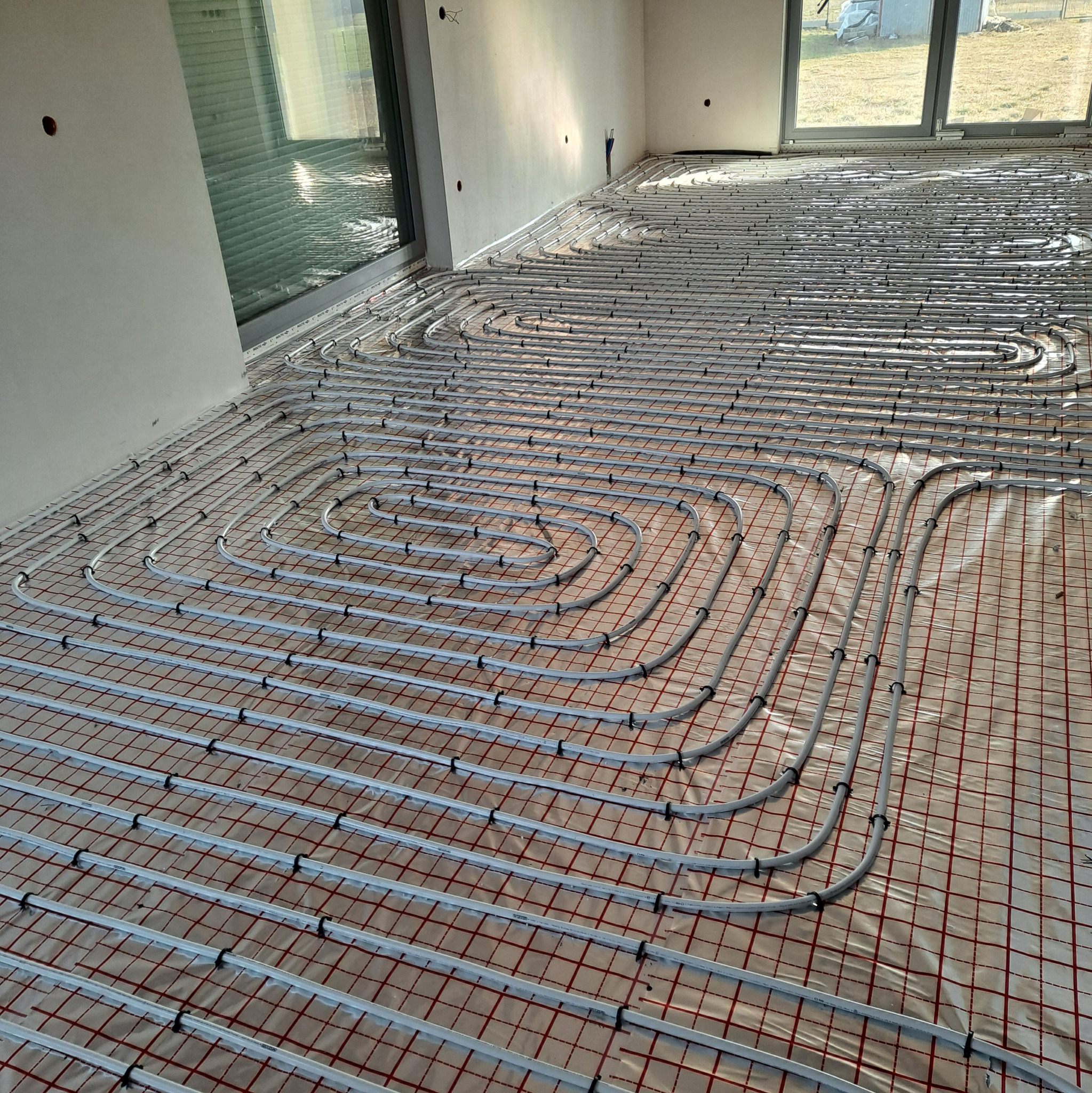 Białe rury ogrzewania podłogowego ułożone w pętle na czerwonej siatce w jasnym pomieszczeniu z dużymi oknami. Etap instalacji przed wylaniem wylewki.