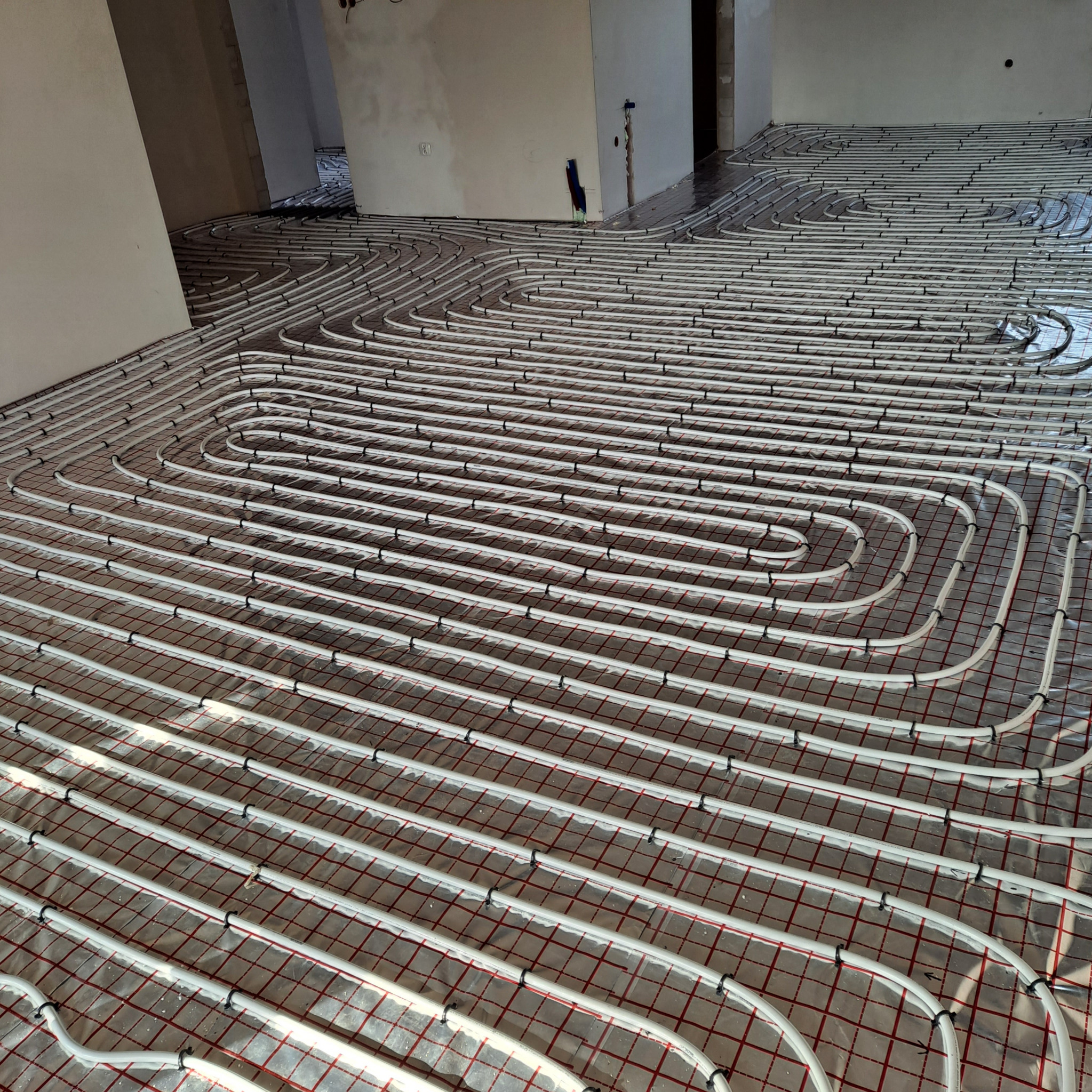 Białe rury ogrzewania podłogowego ułożone spiralnie na folii z czerwoną siatką w surowym wnętrzu. Widoczne elementy instalacji na ścianie.