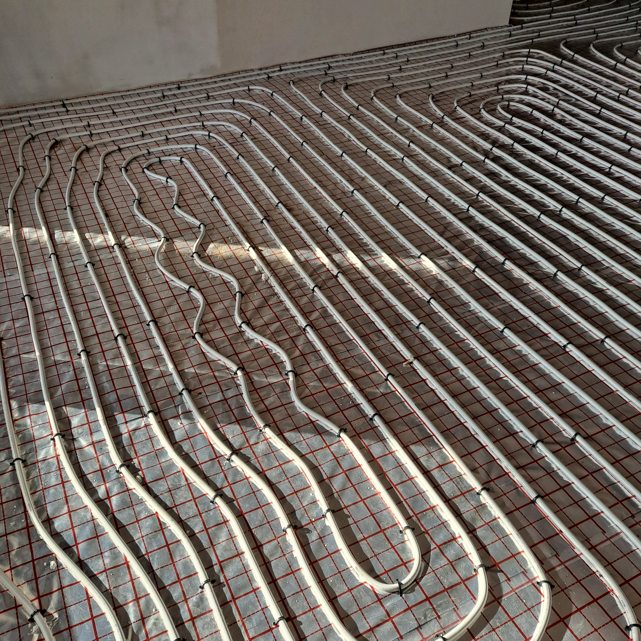 Białe rury ogrzewania podłogowego, równolegle ułożone i przymocowane do czerwonej siatki na folii izolacyjnej, widoczne czarne uchwyty mocujące.