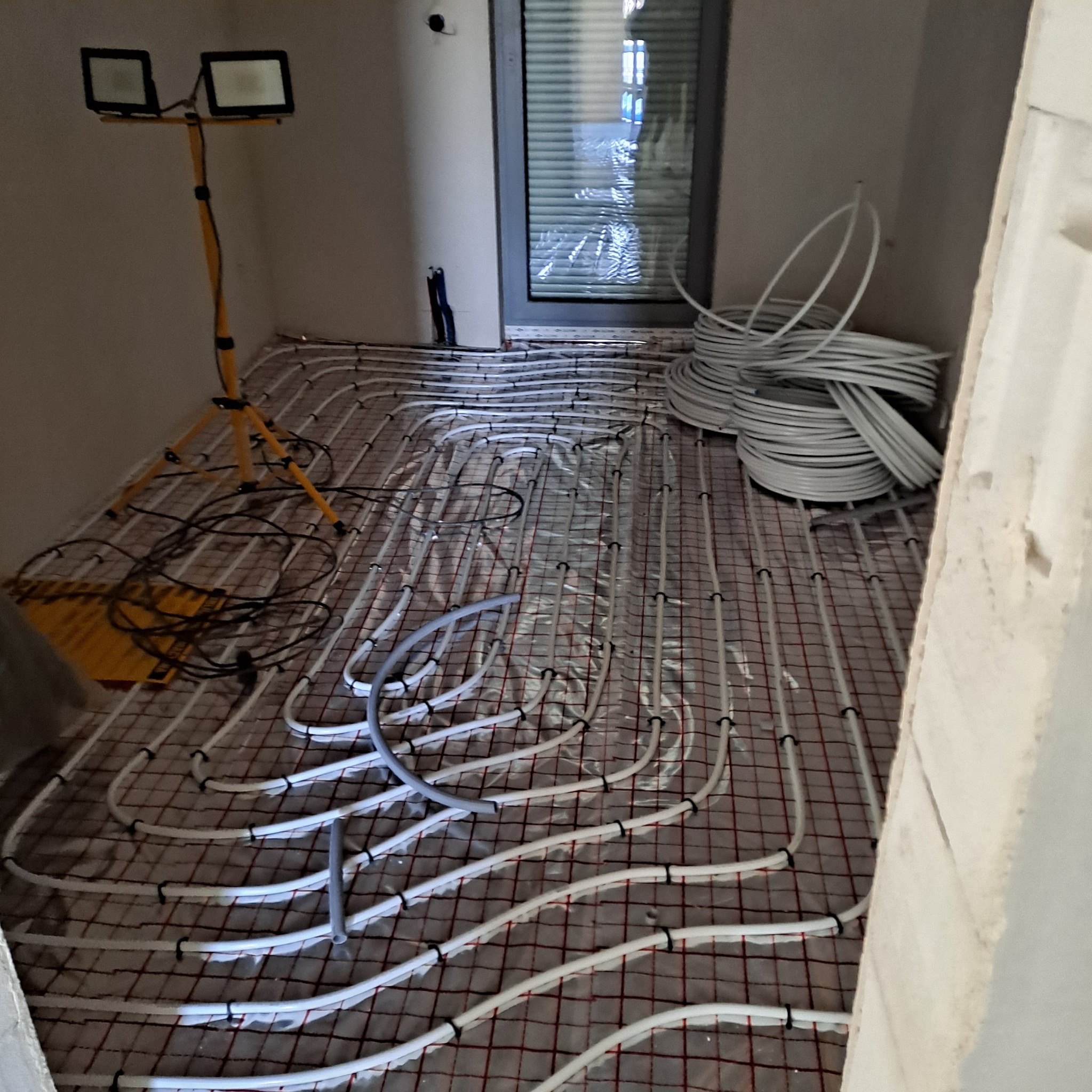 Układanie ogrzewania podłogowego w Władysławowie: białe rurki na siatce, lampa budowlana. Widok na instalację przed wylaniem posadzki, zwoje rur w tle.