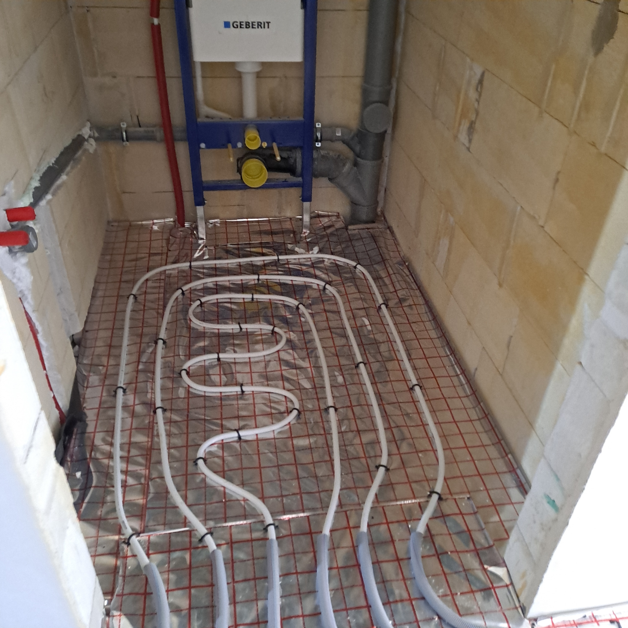 Białe rury ogrzewania podłogowego ułożone na folii w łazience z widoczną instalacją Geberit. Surowe ściany z betonu komórkowego.