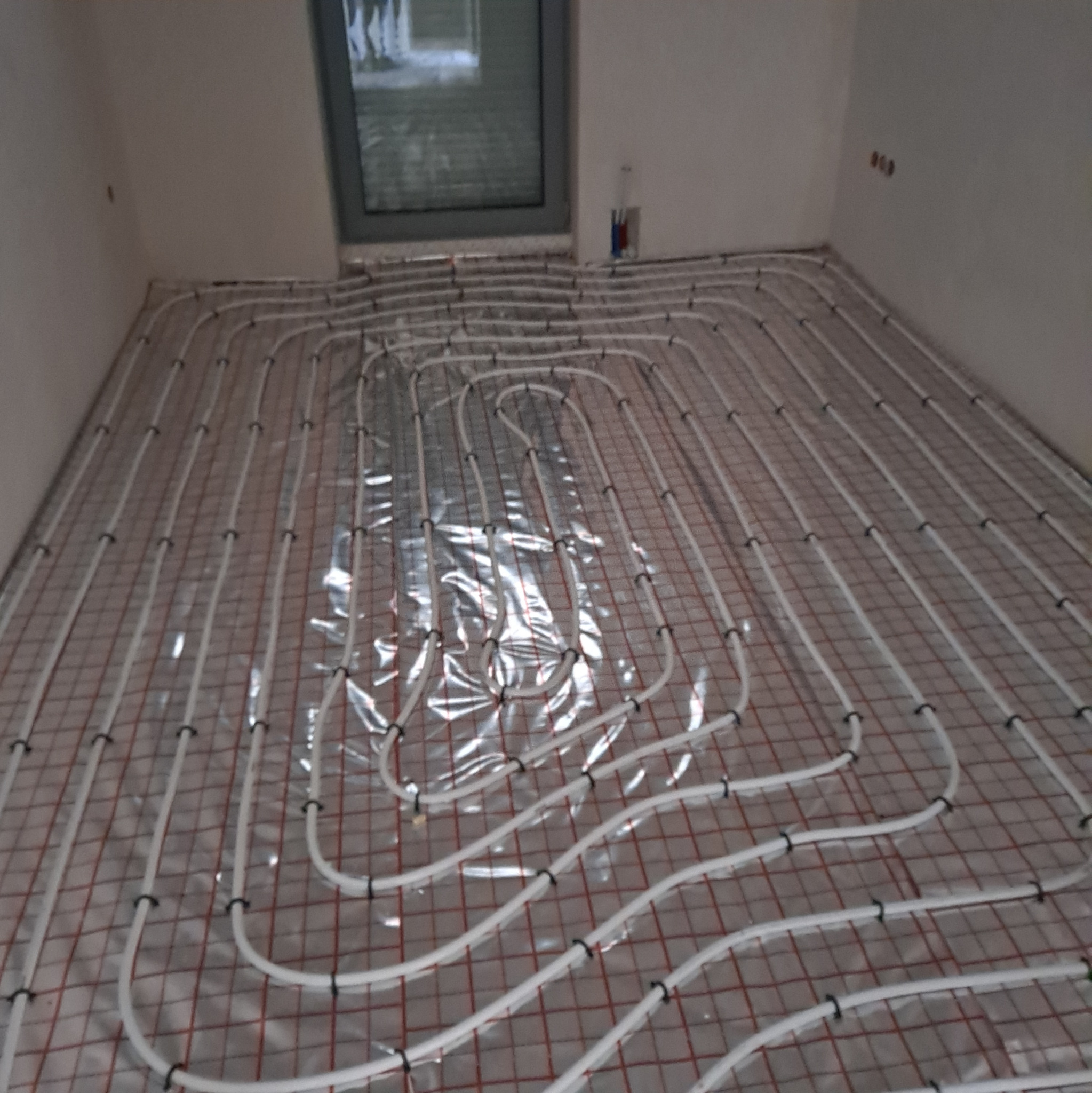 Białe rury ogrzewania podłogowego ułożone na folii z siatką w jasnym pomieszczeniu. Widoczne okno i fragment ściany z instalacją elektryczną.