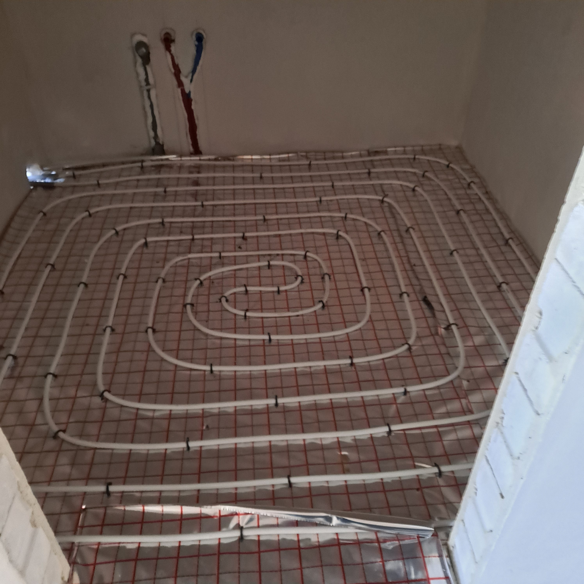 Białe rury ogrzewania podłogowego ułożone spiralnie na folii z czerwoną siatką w surowym wnętrzu. Widoczne wystające rury instalacji.