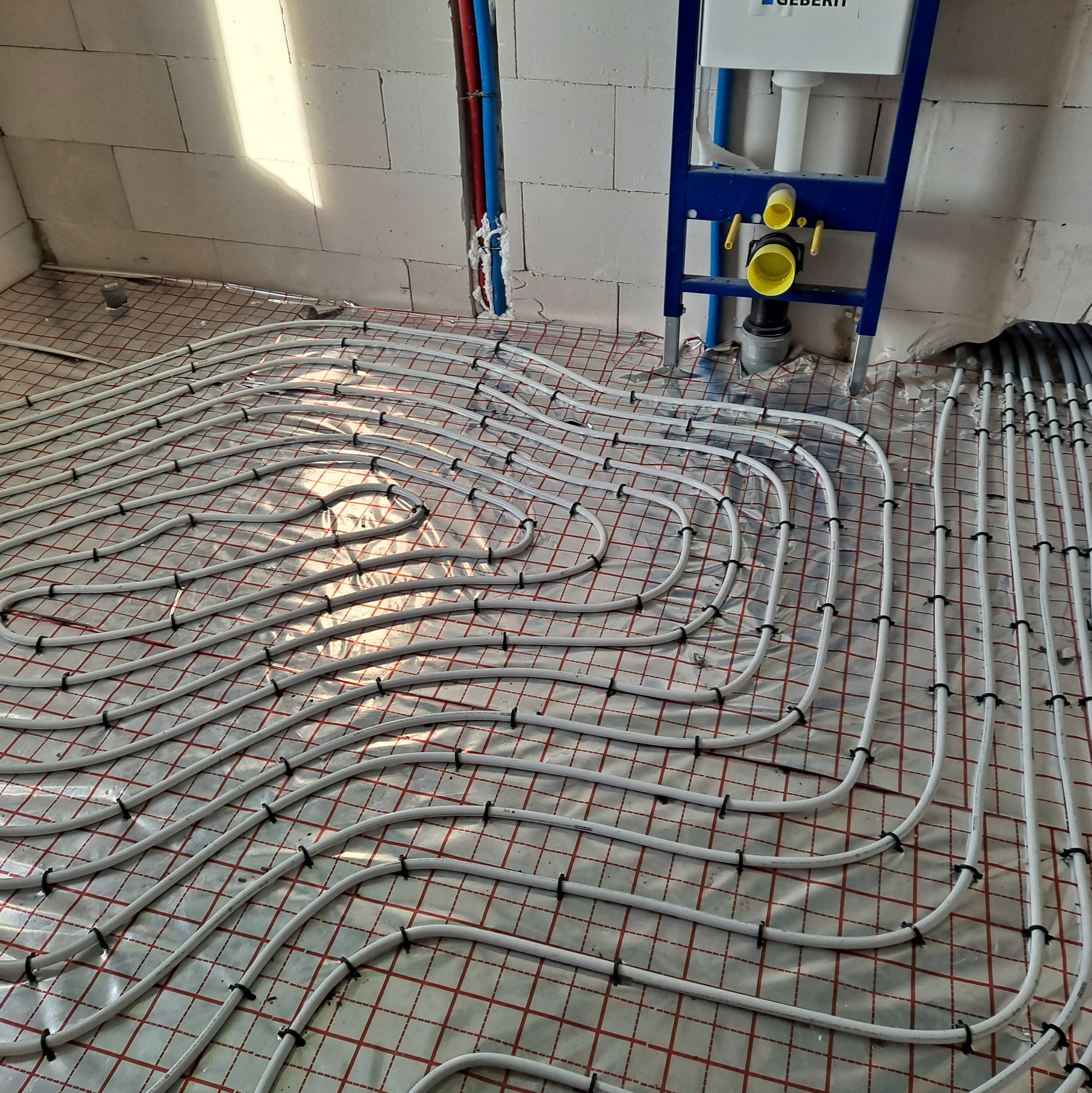 Białe rury ogrzewania podłogowego ułożone na folii z czerwoną siatką, przymocowane czarnymi klipsami. W tle ściana z białego betonu komórkowego i instalacja sanitarna.