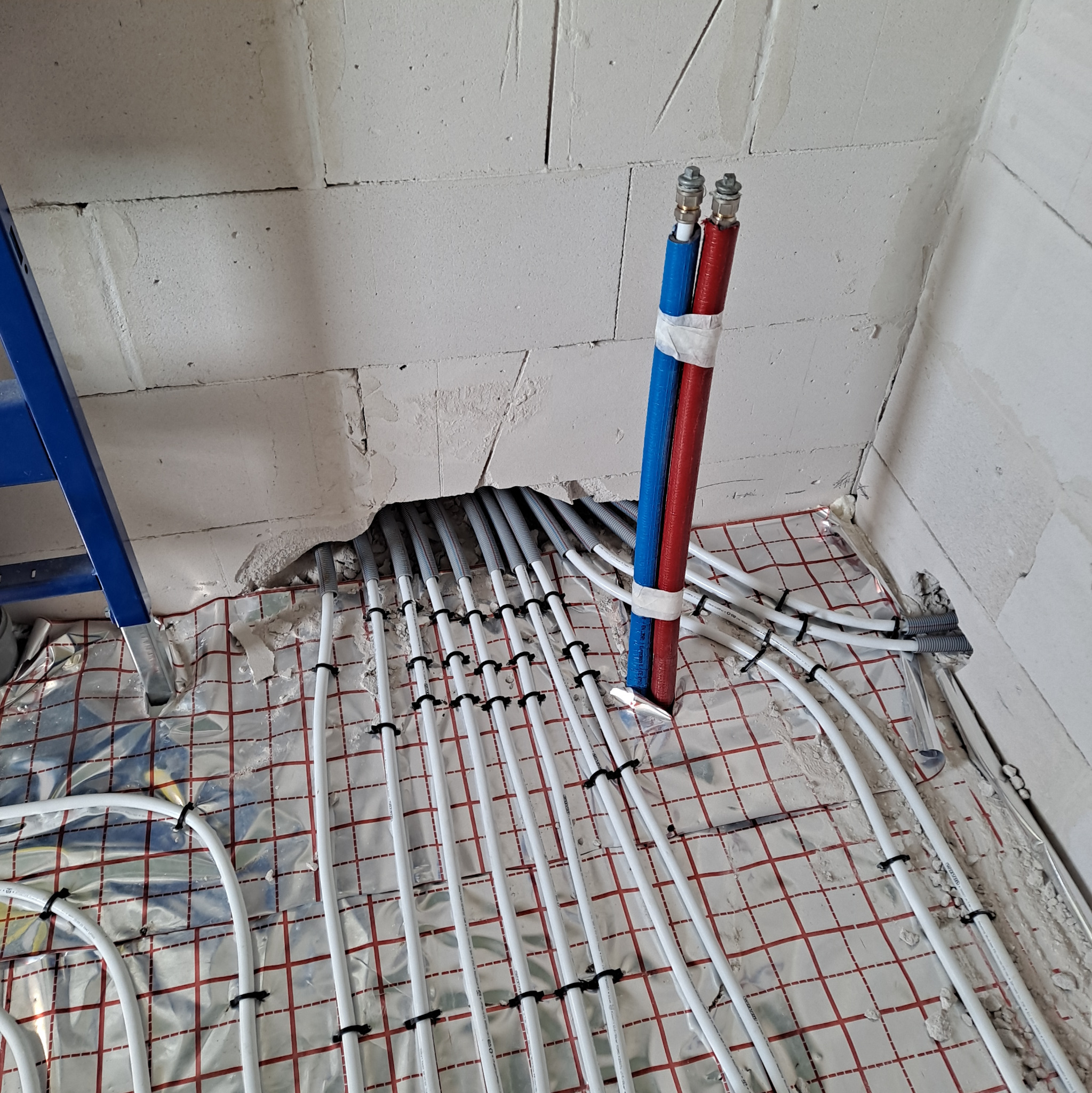 Białe rury ogrzewania podłogowego, mocowane czarnymi uchwytami do folii z czerwoną siatką, wystające z dziury w ścianie z bloków, zasilanie rurami czerwono-niebieskimi.