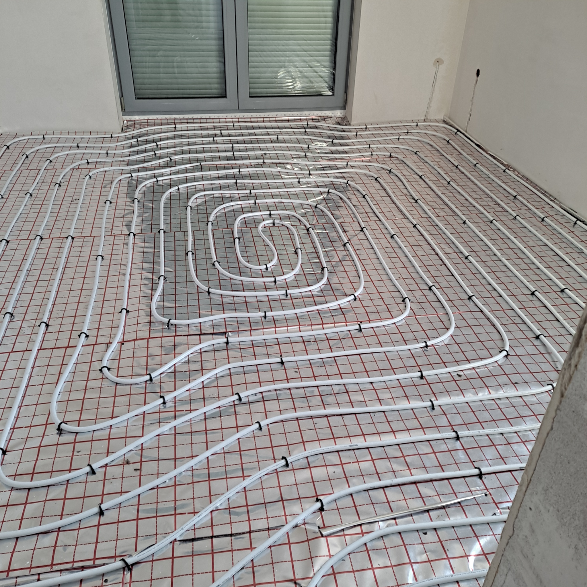 Białe rury ogrzewania podłogowego ułożone na folii z czerwoną siatką, zamocowane czarnymi klipsami, widok z góry, okno w tle.