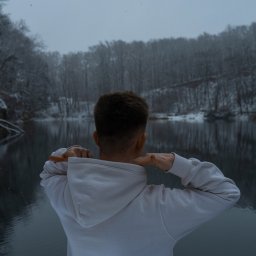 Wilczewski Forografia - Mężczyzna w białej bluzie z kapturem, tyłem do obiektywu, na tle zimowego krajobrazu z jeziorem i ośnieżonym lasem. Zimowa aura, pochmurne niebo.