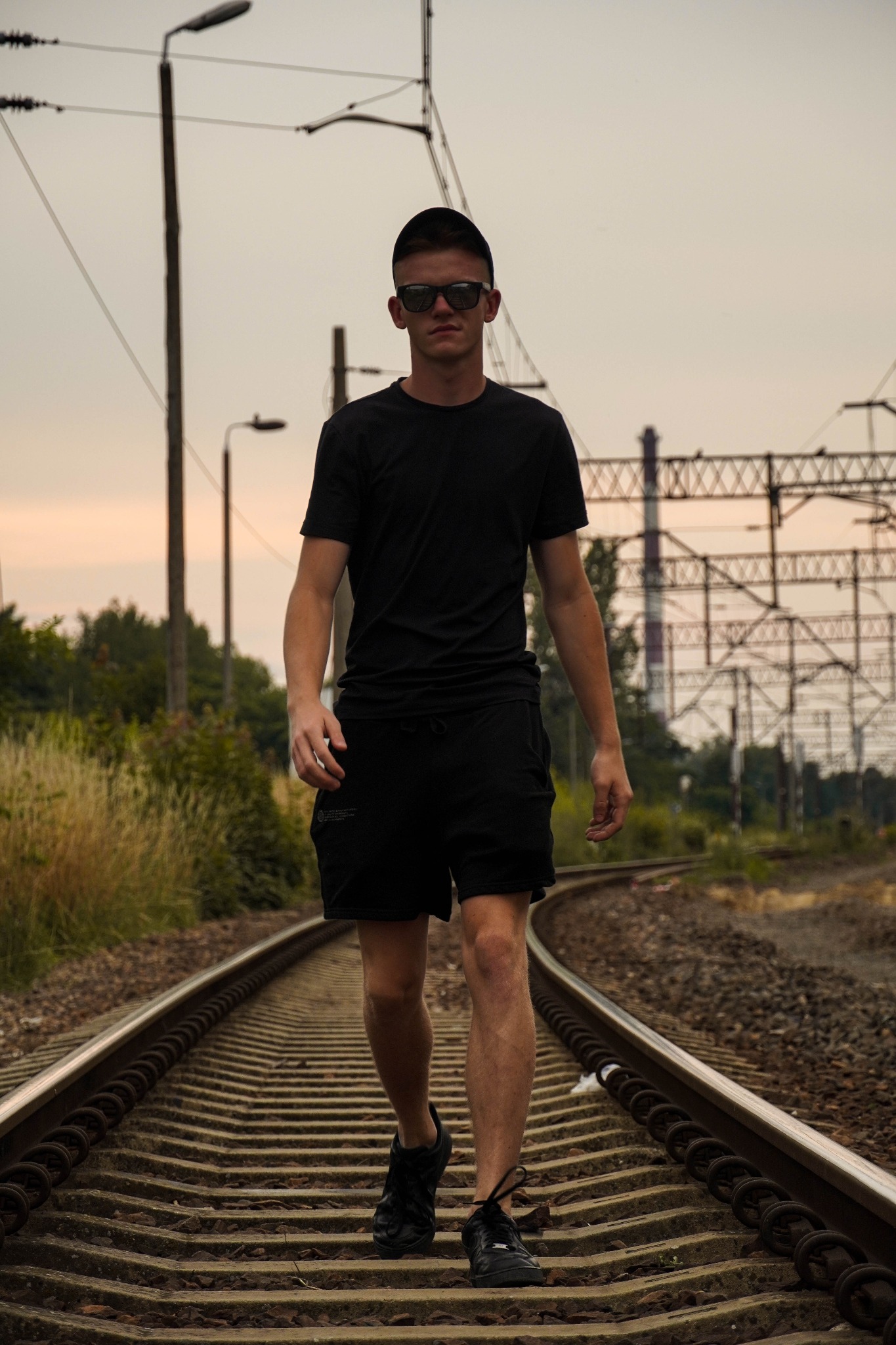 Mężczyzna w czarnym ubraniu i okularach przeciwsłonecznych idzie po torach kolejowych w kierunku obiektywu, z widocznymi słupami i trakcją kolejową w tle.