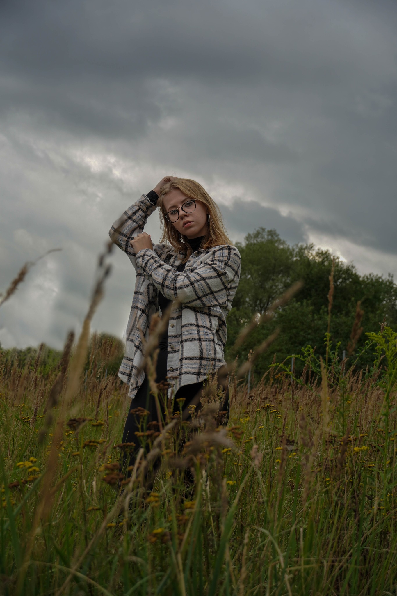 Portret kobiety w okularach, stojącej na łące w pochmurny dzień. Ubrana w czarny golf i flanelową koszulę w kratę. Delikatne, naturalne światło.
