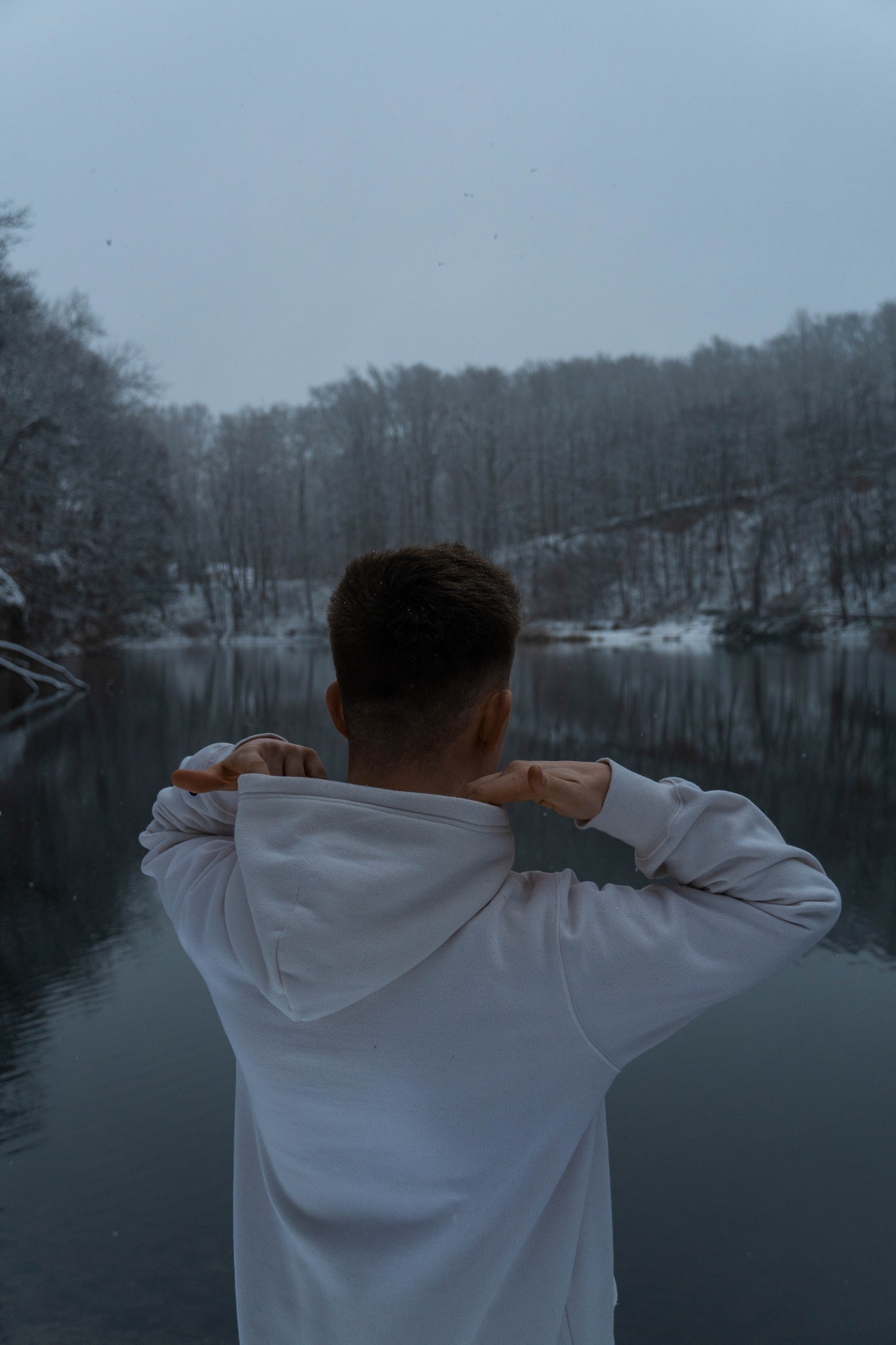 Mężczyzna w białej bluzie z kapturem, tyłem do obiektywu, na tle zimowego krajobrazu z jeziorem i ośnieżonym lasem. Zimowa aura, pochmurne niebo.