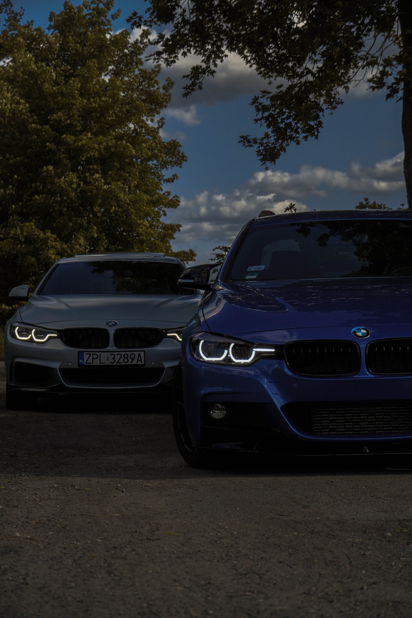 Dwa samochody BMW: błękitny i srebrny, zaparkowane na żwirowej drodze z widocznymi światłami do jazdy dziennej, drzewami i pochmurnym niebem w tle.