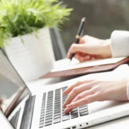 Kobieta pisze ręcznie w notatniku i jednocześnie korzysta z laptopa; roślina doniczkowa w tle, jasne oświetlenie, praca biurowa.