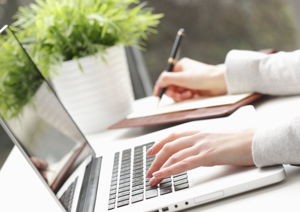 Kobieta pisze ręcznie w notatniku i jednocześnie korzysta z laptopa; roślina doniczkowa w tle, jasne oświetlenie, praca biurowa.