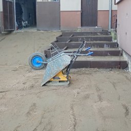 Olekbruk - Przygotowany teren pod kostkę brukową z ubijarką i schodami w tle. Widoczne sznurki wyznaczające obszar. Etap prac przed ułożeniem kostki.