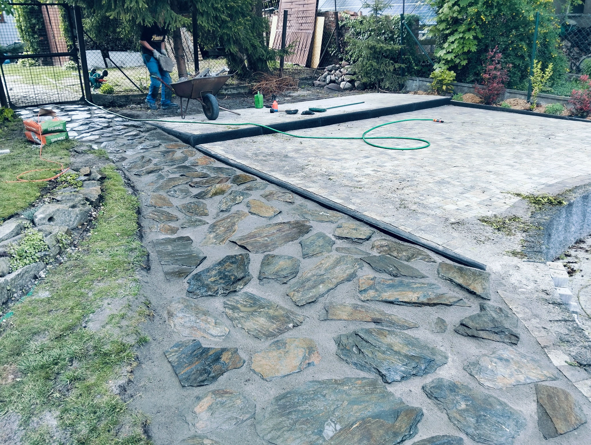 Fragment ogrodu z chodnikiem z nieregularnych kamieni, obok plac z kostki brukowej. W tle osoba z taczką i narzędzia.