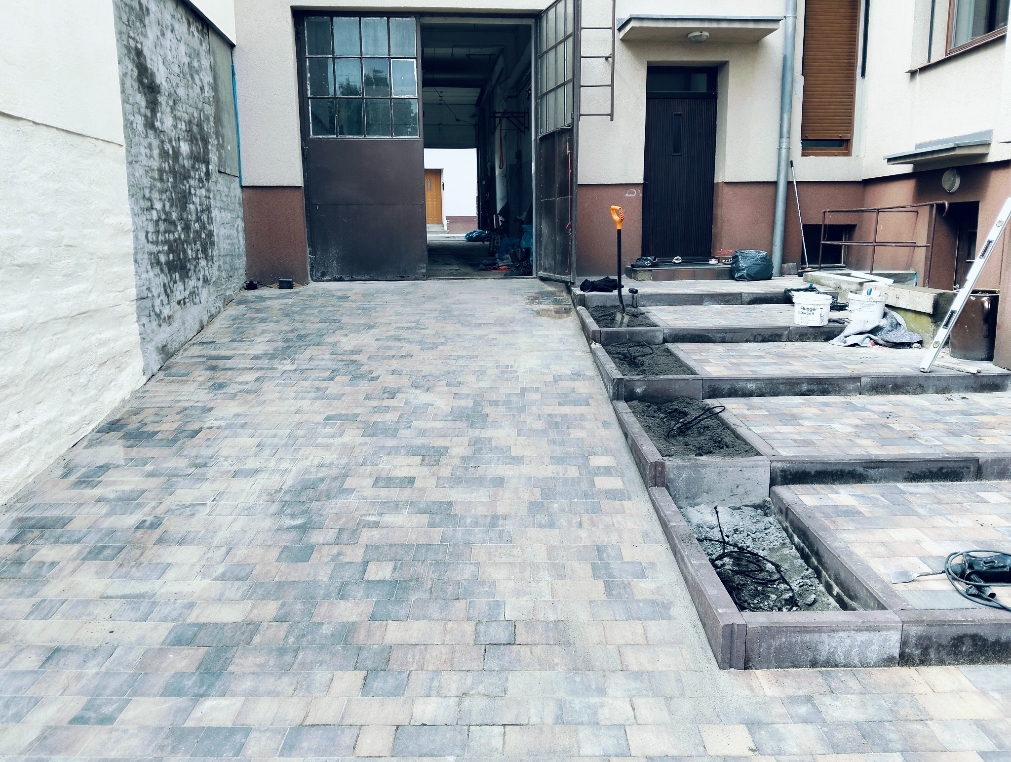 Ułożona kostka brukowa na podjeździe, częściowo w trakcie prac. Widoczne narzędzia i przygotowane fragmenty pod kostkę. W tle budynek z otwartą bramą garażową.