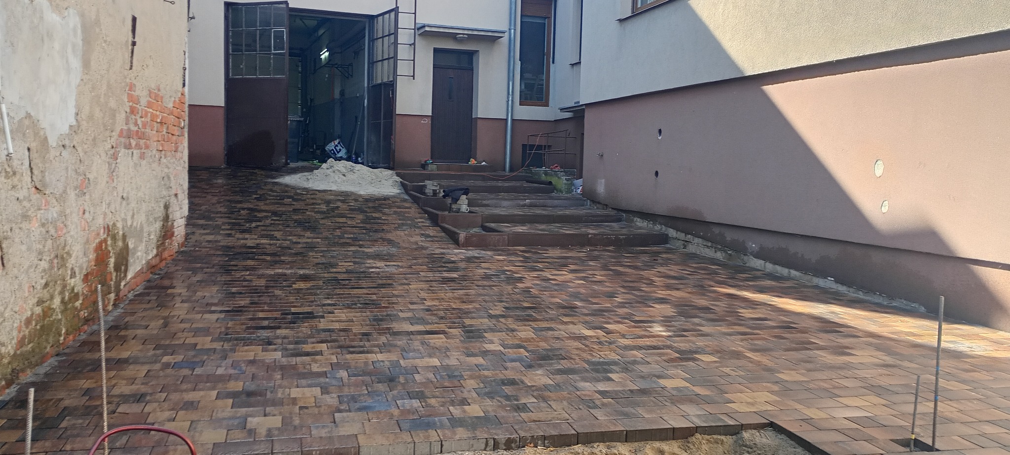 Ułożona kostka brukowa w odcieniach brązu prowadzi do schodów i budynku. Na górze hałda piasku. Część kostki jeszcze nieułożona. Widoczne pręty zbrojeniowe.