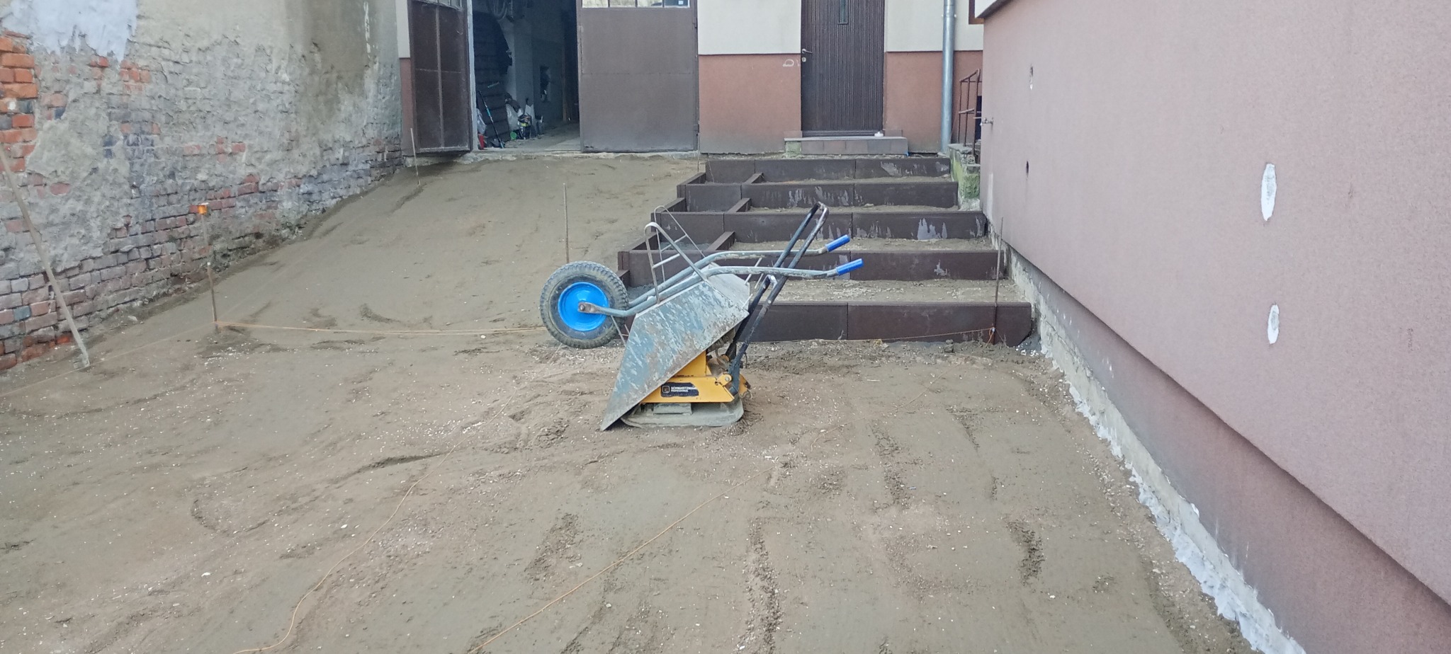 Przygotowany teren pod kostkę brukową z ubijarką i schodami w tle. Widoczne sznurki wyznaczające obszar. Etap prac przed ułożeniem kostki.