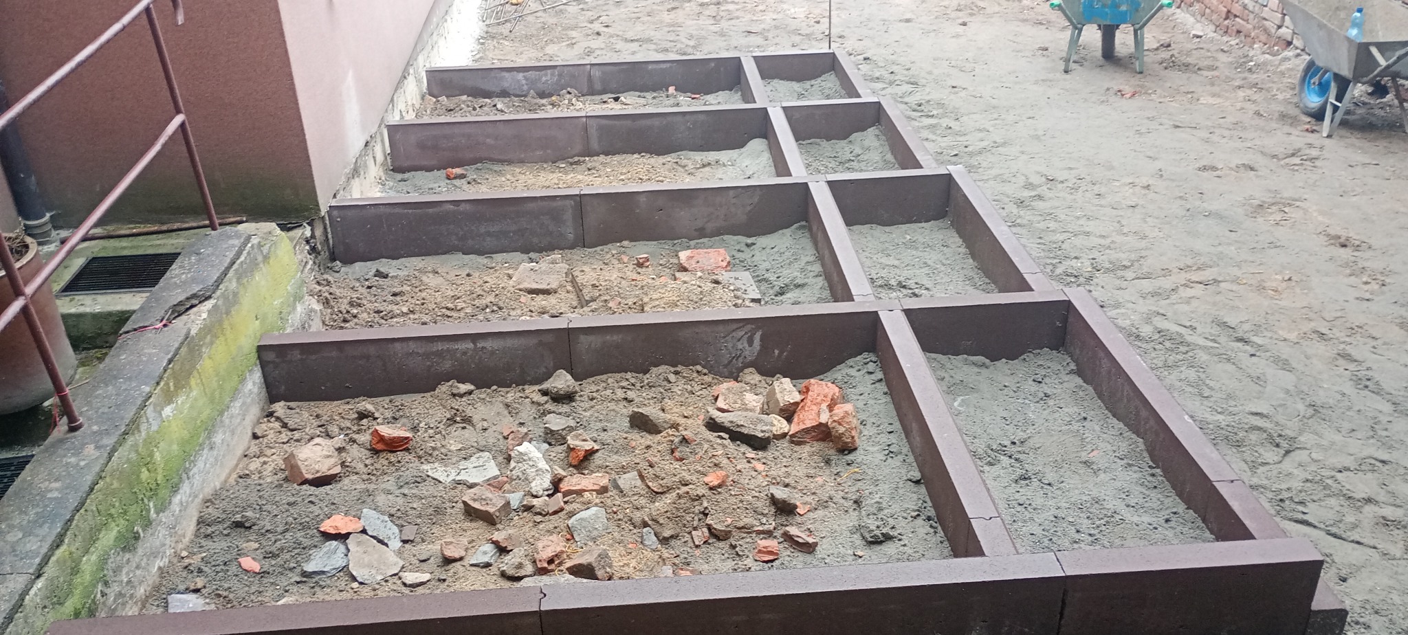 Przygotowanie podłoża pod taras z kostki: widoczne ramy z obrzeży, wypełnione częściowo piaskiem i gruzem. W tle widać taczki i fragment budynku.