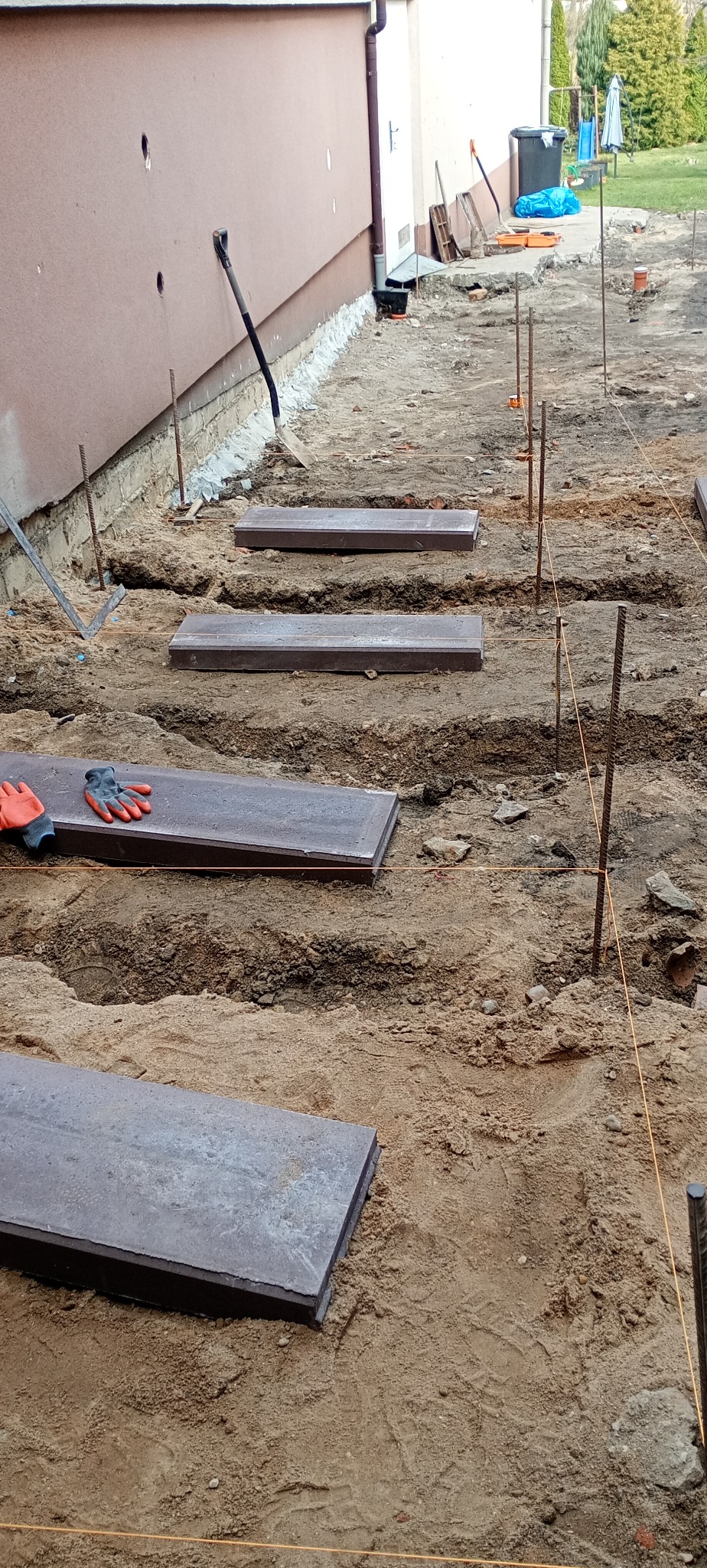 Etap przygotowania podłoża pod kostkę brukową, widoczne płyty ułożone w wykopach, wyznaczona niwelacja terenu, rękawice robocze.