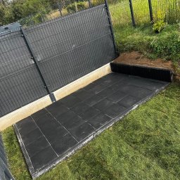 Plantacja Choinek - Wykonana podbudowa z czarnych płyt gumowych w ogrodzie, obok trawnik. Podwyższona konstrukcja z siatki panelowej w tle. Wykopana ziemia wzdłuż krawędzi.