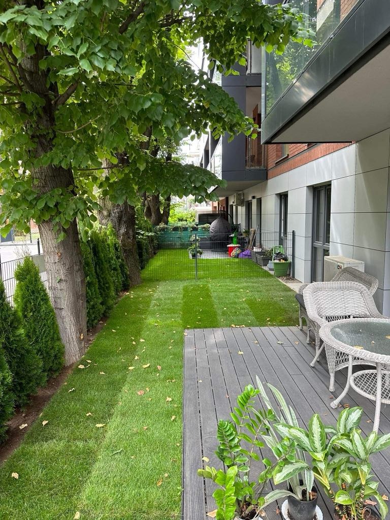 Świeżo skoszony trawnik z wyraźnymi pasami na tle nowoczesnego budynku z balkonami, obok drewniany taras z roślinami doniczkowymi i meblami ogrodowymi.
