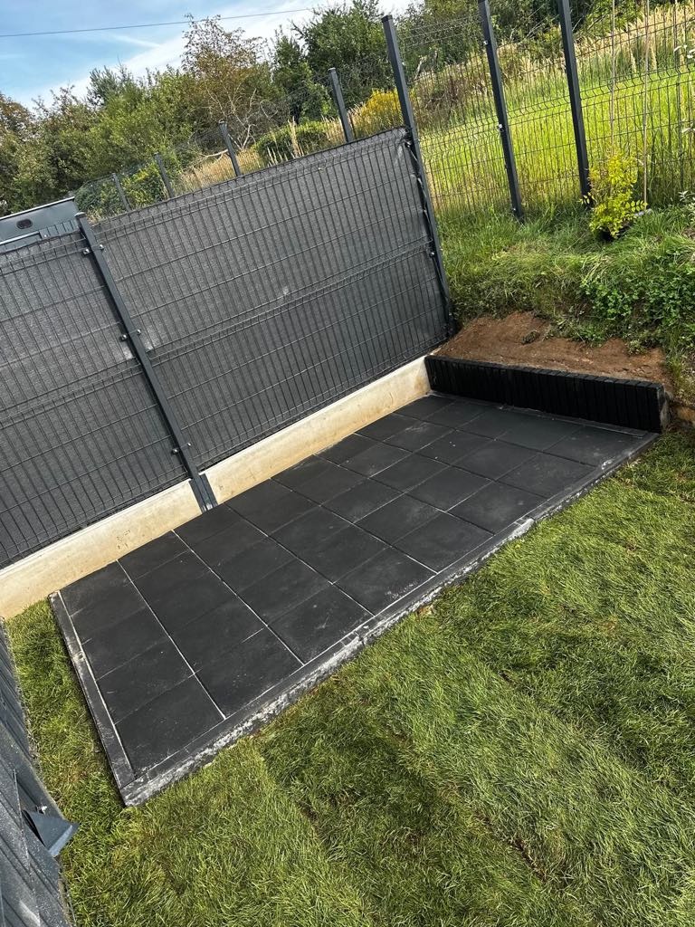 Wykonana podbudowa z czarnych płyt gumowych w ogrodzie, obok trawnik. Podwyższona konstrukcja z siatki panelowej w tle. Wykopana ziemia wzdłuż krawędzi.