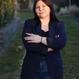 Portret kobiety w granatowej marynarce, skrzyżowane ręce, dżinsy, na tle zieleni ogrodu o zmierzchu. Delikatny uśmiech, naturalne światło podkreśla rysy twarzy.