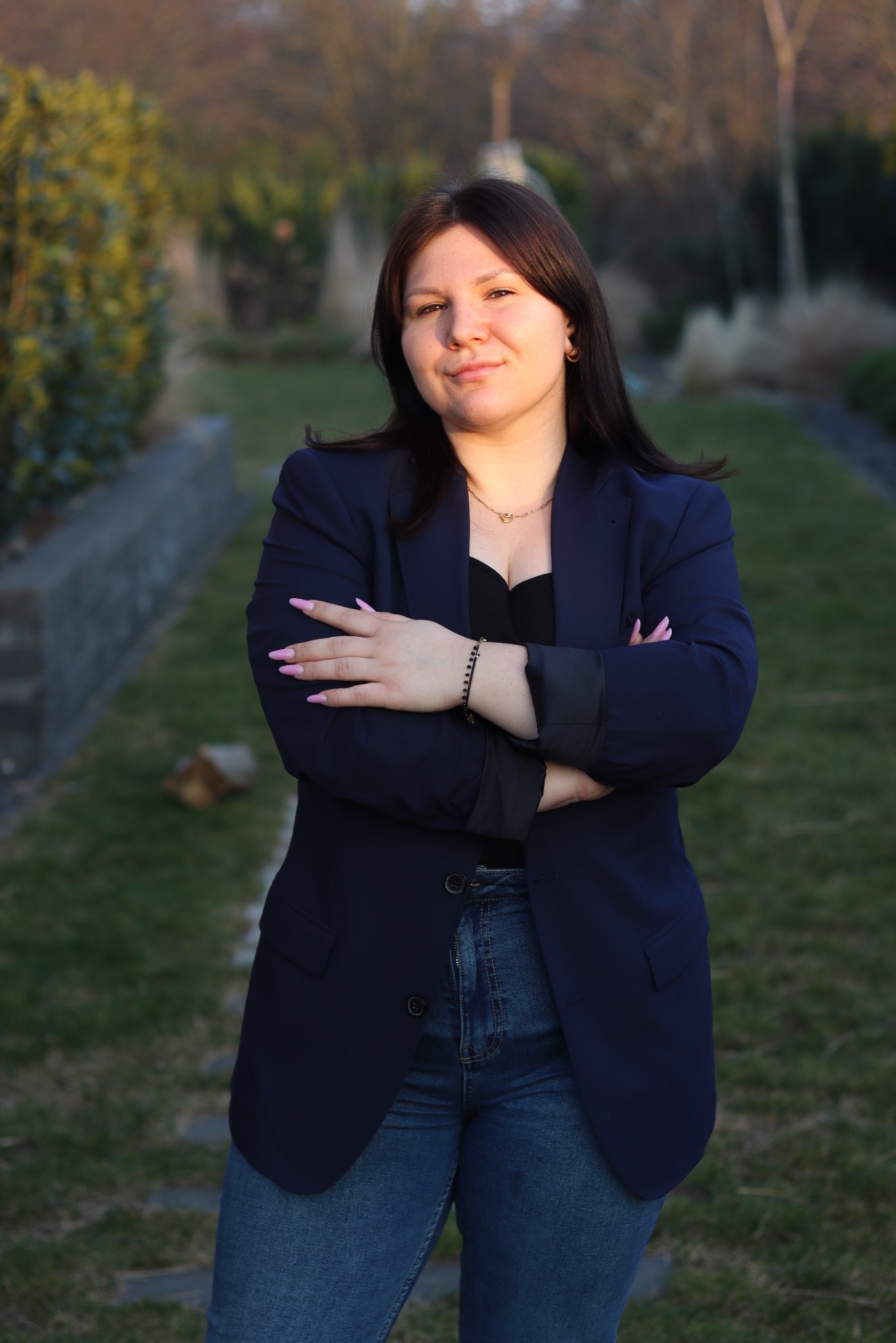 Portret kobiety w granatowej marynarce, skrzyżowane ręce, dżinsy, na tle zieleni ogrodu o zmierzchu. Delikatny uśmiech, naturalne światło podkreśla rysy twarzy.