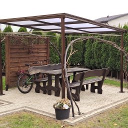 Denys Kokoshko Service - Zadaszona altana z drewnianym stołem i ławkami na wybrukowanym patio, otoczona tujami. Na pierwszym planie siedzący kot. Z boku oparta o altanę zielona rower.