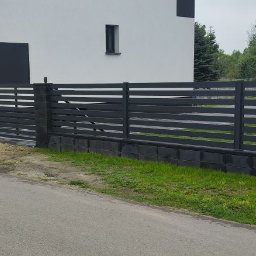 Denys Kokoshko Service - Nowoczesne, poziome ogrodzenie metalowe w kolorze grafitowym na podmurówce z betonowych bloczków, otaczające posesję z białym domem w tle.