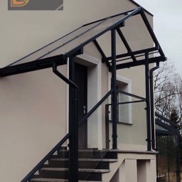 Denys Kokoshko Service - Zadaszenie nad schodami wejściowymi z czarną, metalową konstrukcją i przezroczystym wypełnieniem. Dodatkowo balustrady ze szkła i metalu. Widok z dołu.