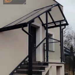 Zadaszenie nad schodami wejściowymi z czarną, metalową konstrukcją i przezroczystym wypełnieniem. Dodatkowo balustrady ze szkła i metalu. Widok z dołu.