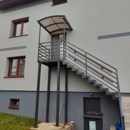 Denys Kokoshko Service - Metalowe schody zewnętrzne z poręczą i zadaszeniem prowadzące do drzwi budynku. Widoczne okna i śmietnik. Szara kolorystyka.