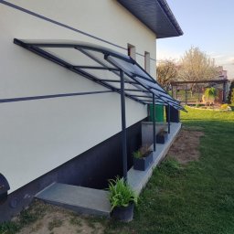 Denys Kokoshko Service - Zadaszenie nad wejściem do piwnicy w domu jednorodzinnym. Konstrukcja stalowa w kolorze grafitowym, wypełnienie z przezroczystego tworzywa. Widok z boku, trawnik, drzewa w tle.