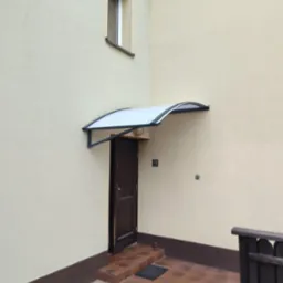 Zadaszenie nad brązowymi drzwiami wejściowymi do domu z beżową elewacją. Widoczny fragment ogrodzenia i okno na piętrze. Kąt prosty budynku.