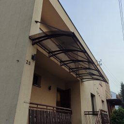 Denys Kokoshko Service - Zadaszenie nad wejściem do budynku z numerem 25. Konstrukcja metalowa z przezroczystym wypełnieniem, widoczne elementy mocujące do ściany. Ujęcie z dołu, widok na elewację.