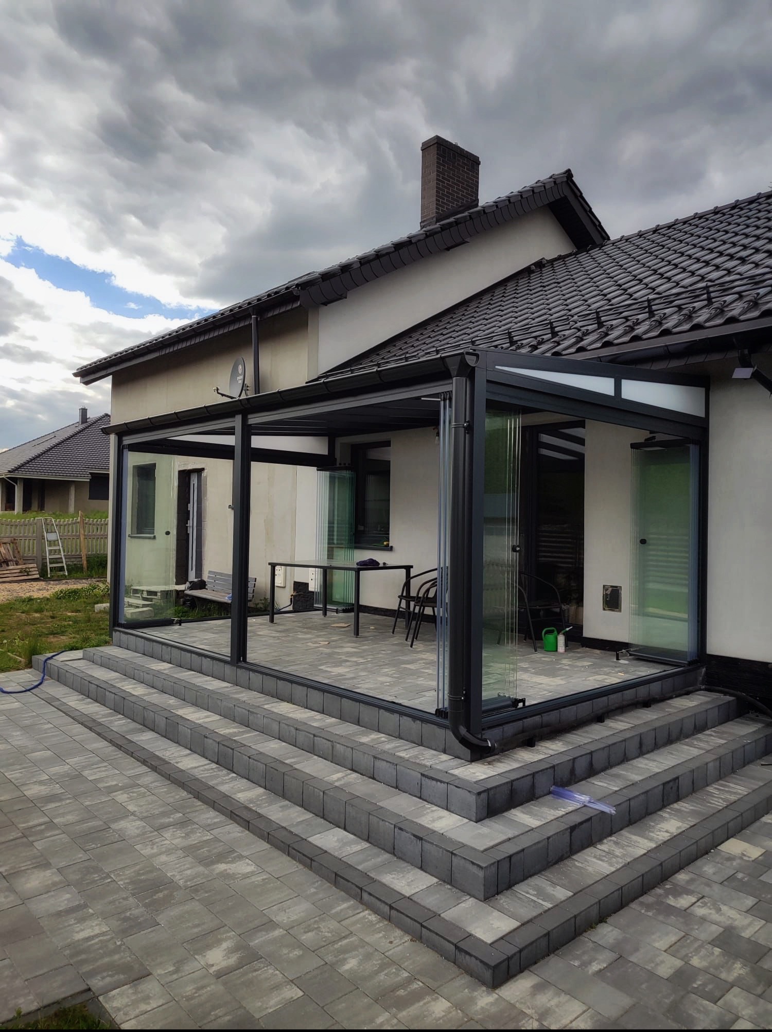 Nowoczesna zabudowa tarasu z przesuwnymi szklanymi panelami, widoczna z zewnątrz na tle domu z ciemnym dachem i brukowanymi schodami wejściowymi.