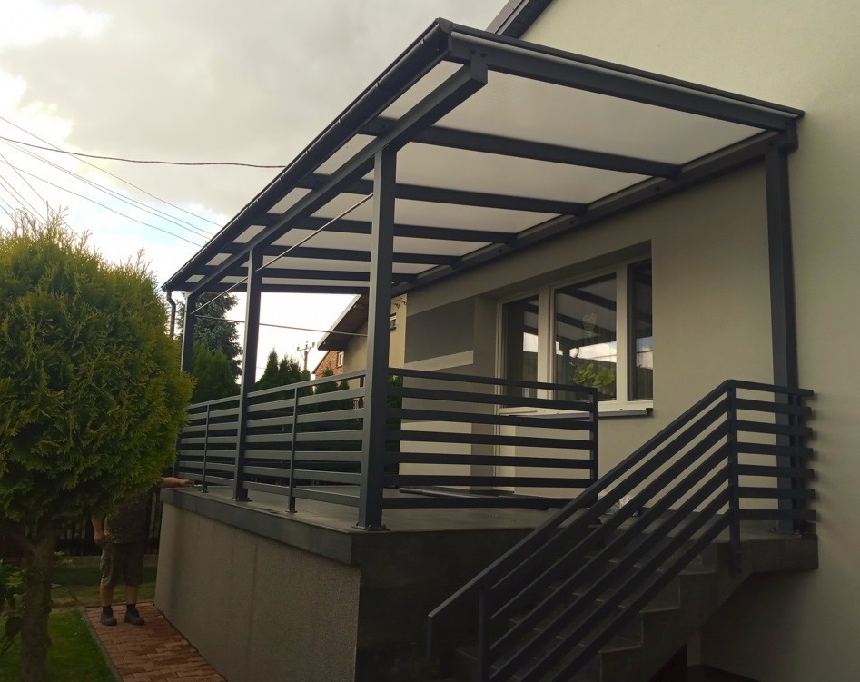 Zadaszony taras z metalową balustradą w nowoczesnym stylu, z widocznymi schodami i roślinnością w tle. Elementy konstrukcyjne w kolorze grafitowym.