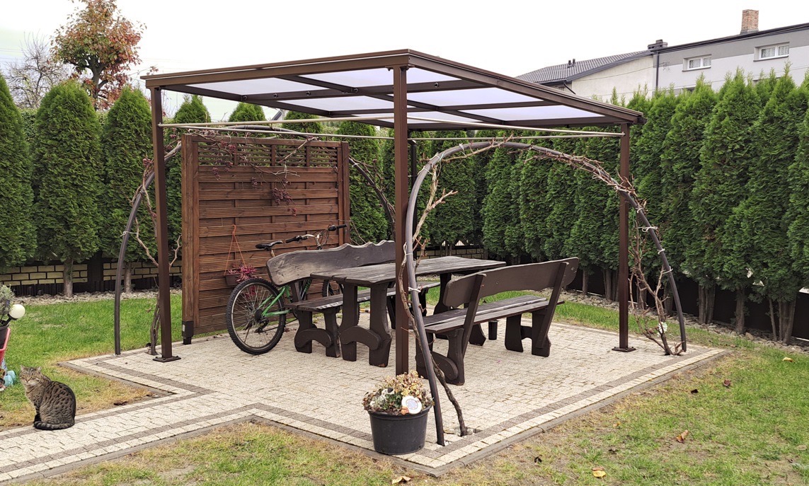 Zadaszona altana z drewnianym stołem i ławkami na wybrukowanym patio, otoczona tujami. Na pierwszym planie siedzący kot. Z boku oparta o altanę zielona rower.