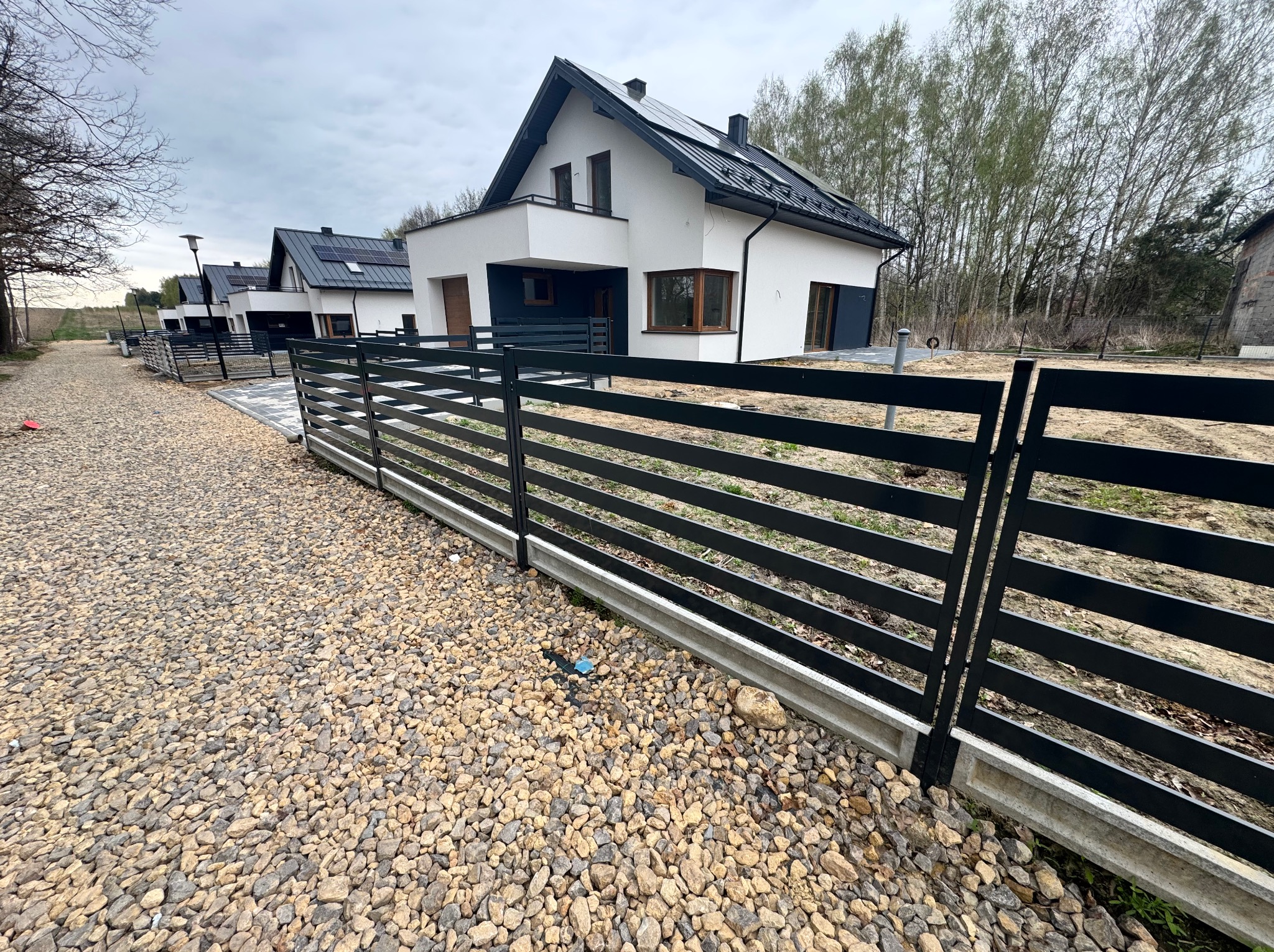 Nowoczesne ogrodzenie panelowe w kolorze czarnym, z betonową podmurówką, na tle szeregu domów z panelami słonecznymi w słoneczny dzień. Widok z bliska na żwirowe podłoże.