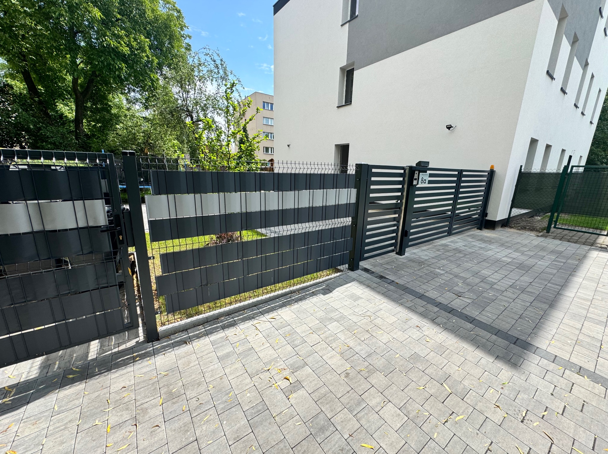 Nowoczesna brama wjazdowa i furtka z metalowymi panelami w kolorze grafitu i szarości, na tle jasnego budynku i brukowanej nawierzchni.