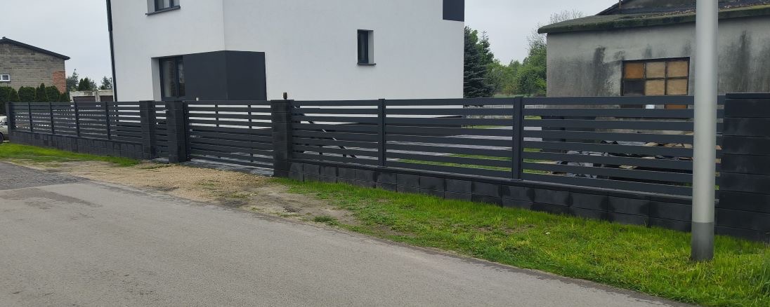 Nowoczesne, poziome ogrodzenie metalowe w kolorze grafitowym na podmurówce z betonowych bloczków, otaczające posesję z białym domem w tle.