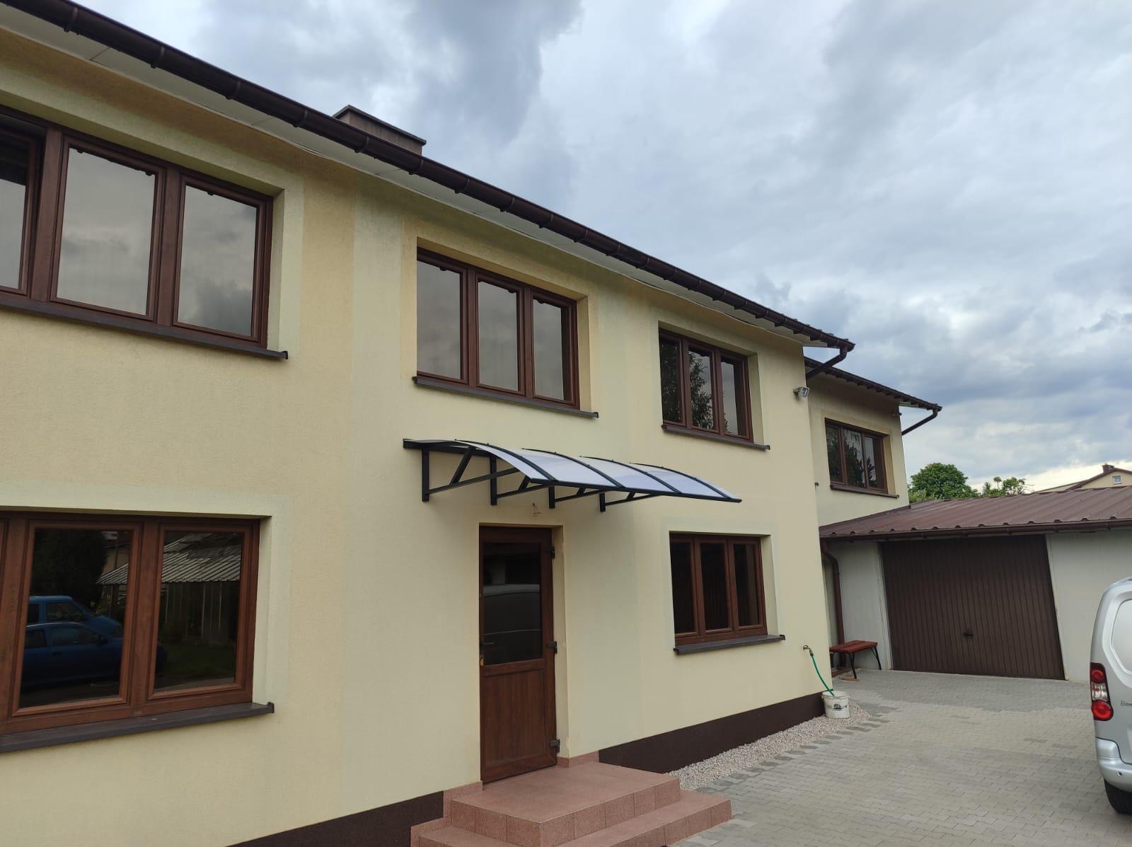 Dom z beżową elewacją i brązowymi oknami. Nad drzwiami wejściowymi zamontowany daszek z metalową konstrukcją i przezroczystym wypełnieniem.