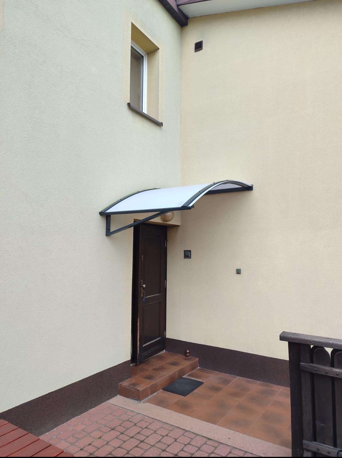 Zadaszenie nad brązowymi drzwiami wejściowymi do domu z beżową elewacją. Widoczny fragment ogrodzenia i okno na piętrze. Kąt prosty budynku.