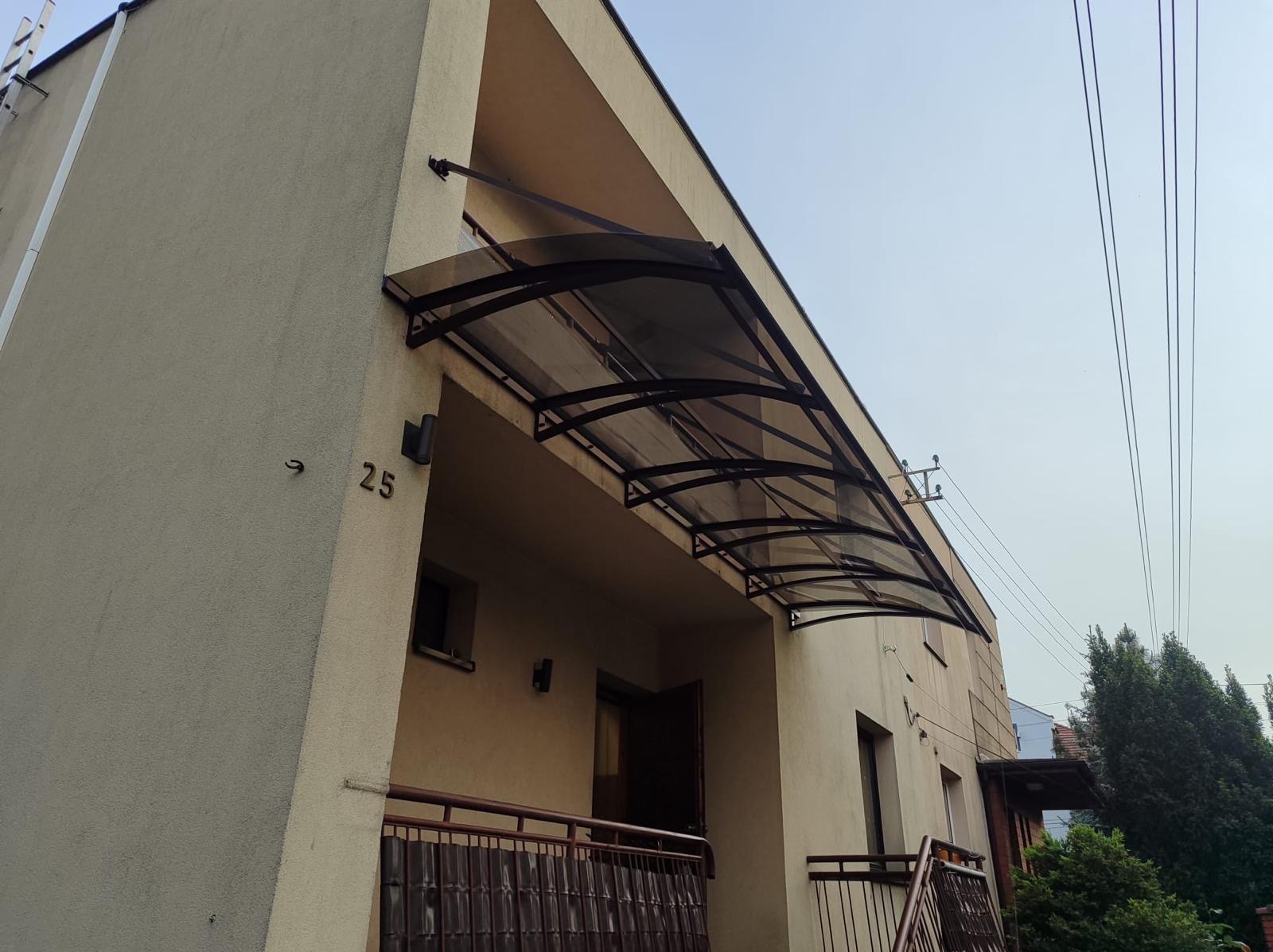Zadaszenie nad wejściem do budynku z numerem 25. Konstrukcja metalowa z przezroczystym wypełnieniem, widoczne elementy mocujące do ściany. Ujęcie z dołu, widok na elewację.