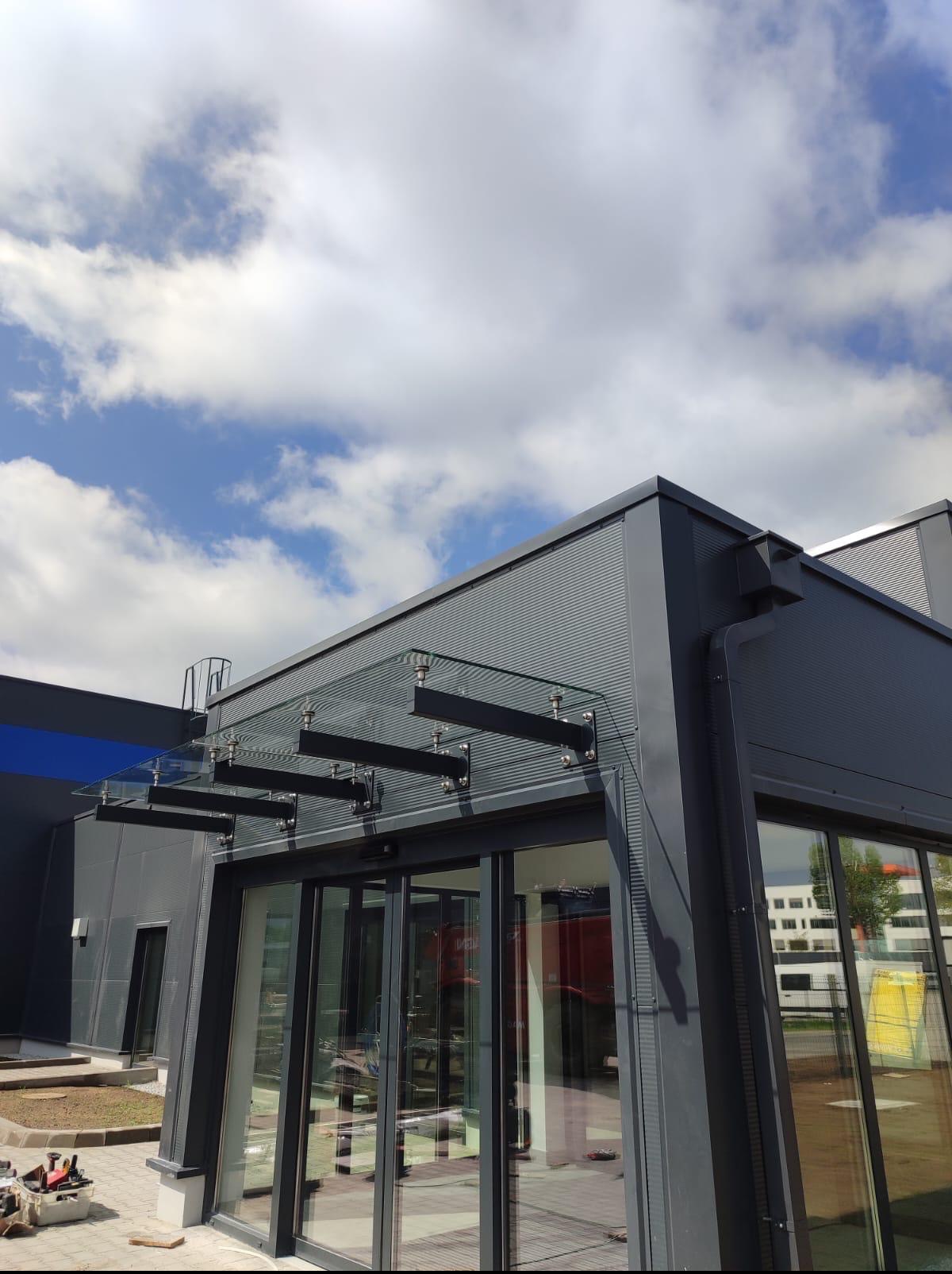 Szklany daszek nad wejściem do budynku o szarej, metalowej elewacji. Widoczne niebo z chmurami. Na pierwszym planie skrzynka z narzędziami.