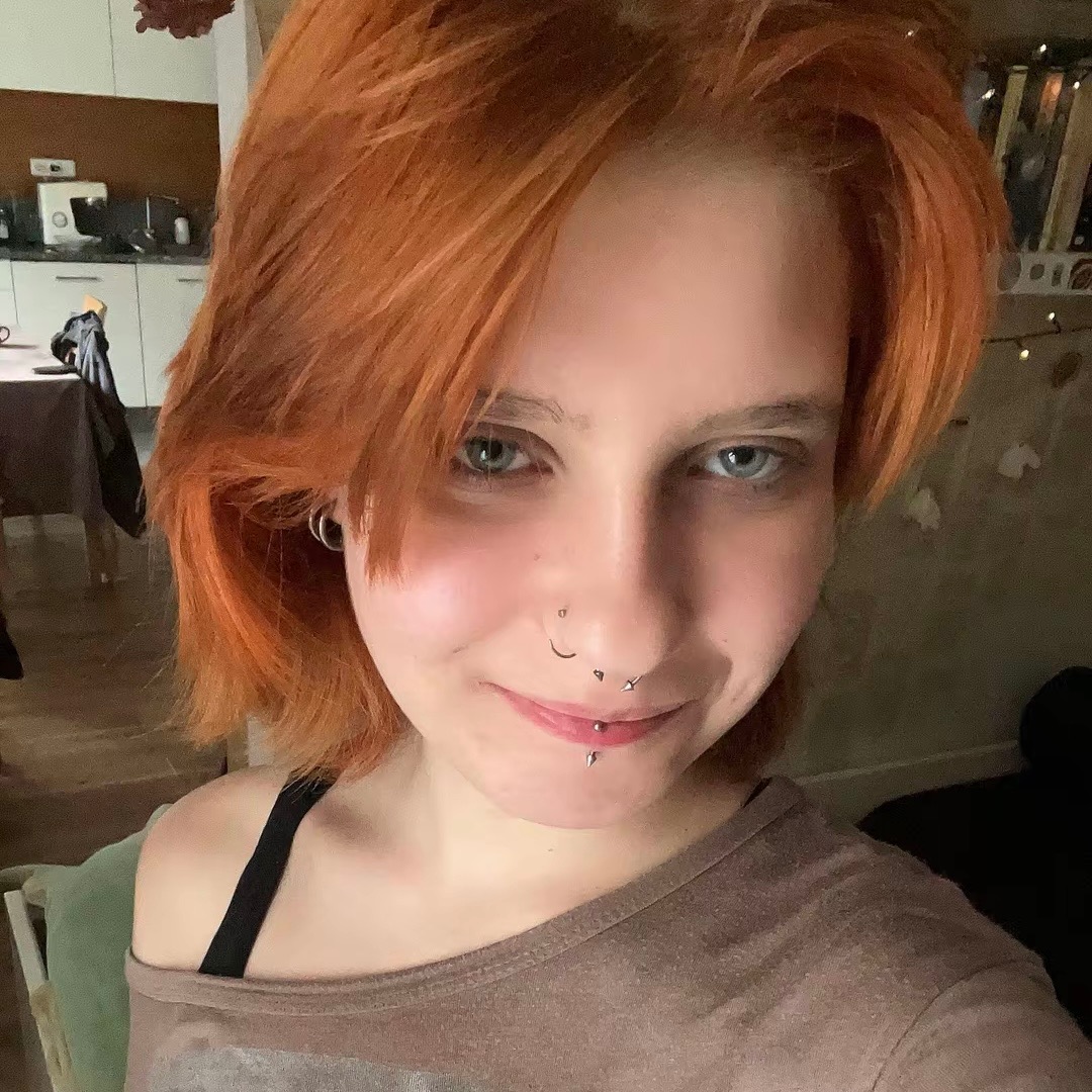 Portret uśmiechniętej kobiety z rudymi włosami i piercingiem na twarzy, w brązowej bluzce na tle wnętrza mieszkania.