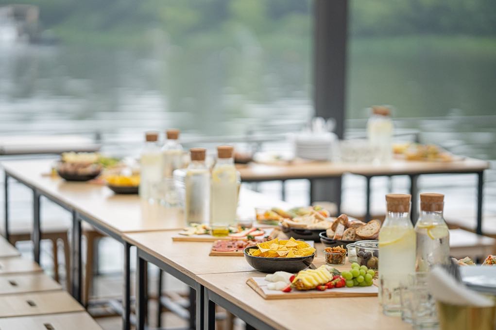 Stół cateringowy z przekąskami i napojami na tle jeziora. Widoczne deski z serami, owocami, pieczywo i lemoniada w szklanych butelkach z korkiem.